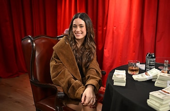 Una mujer de cabello oscuro y blusa negra se sienta a una mesa negra con fichas de póker, billetes, una lata de bebida y un vaso, frente a una cortina roja