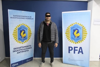 Hombre con rostro censurado, chaqueta gris y pantalón oscuro, de pie entre dos estandartes de la Policía Federal Argentina con logotipos y texto