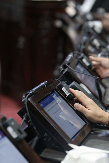 Una mano presiona un botón negro en una consola de votación con pantalla en un hemiciclo legislativo, mostrando parte de un micrófono y texto en la pantalla