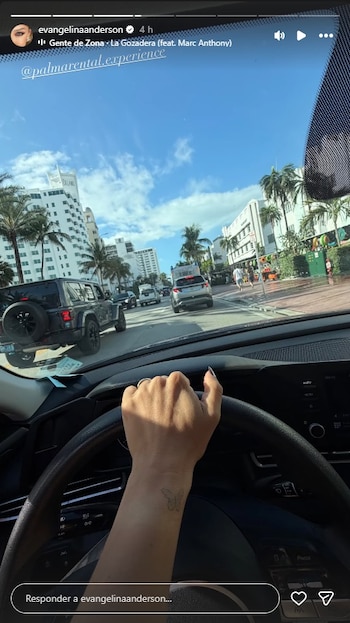 Vista desde un coche en Miami con una mano al volante y tatuaje de mariposa. La calle muestra tráfico, palmeras y edificios bajo un cielo azul