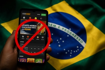 Una mano sostiene un teléfono inteligente que muestra una aplicación de apuestas con un símbolo de prohibición sobre la bandera de Brasil.