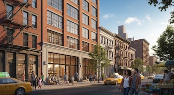 Calle urbana con edificios de ladrillo y fachadas comerciales. Se ven personas caminando, taxis amarillos, un puesto de frutas y árboles bajo un cielo azul claro.