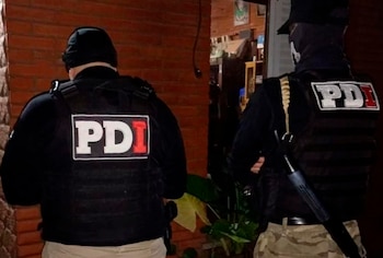Dos agentes de la PDI, de espaldas, con chalecos tácticos negros y logotipos PDI, parados en la entrada de una casa con pared de ladrillos