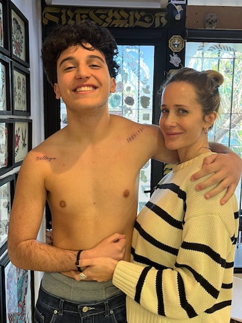 Un joven con el torso descubierto y dos tatuajes en los hombros abraza a una mujer rubia con un suéter a rayas en un estudio