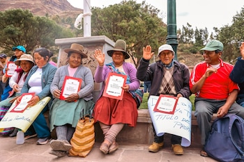 Hombres y mujeres sentados al aire libre sostienen carpetas rojas con documentos oficiales y pancartas de papel manuscritas
