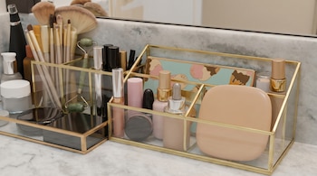 Primer plano de productos cosméticos y herramientas de maquillaje, incluyendo brochas, botellas y compactos, organizados en estantes de vidrio dorados sobre mármol claro.