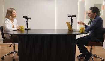 Dra. Giulia Enders sentada a la izquierda de una mesa negra, conversando con un hombre a la derecha. Ambos tienen micrófonos y tazas amarillas, con un portátil abierto