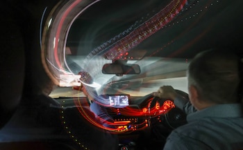 Interior de un automóvil de noche con conductor, pasajero, volante, salpicadero iluminado en rojo y estelas de luz blanca y roja que atraviesan el parabrisas