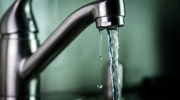 Grifo de acero inoxidable cepillado con un chorro de agua cayendo en hilo continuo y varias gotas de agua suspendidas, fondo desenfocado.