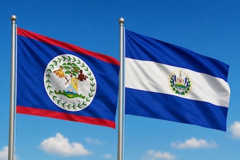 Banderas oficiales de Belice y El Salvador sujetas a mástiles metálicos frente a un cielo azul con nubes blancas.