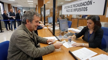 Hombre sonriente con billetes, mujer estampando un documento, mostrador con barrera, cartel "Oficina de Recaudación - Pago de Impuestos", personas y computadoras.