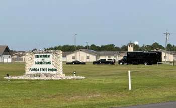 Un cartel de piedra con el texto Department of Corrections Florida State Prison en un terreno de hierba con vehículos y edificios al fondo
