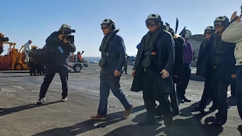 Javier Milei y otros individuos con cascos y chalecos salvavidas caminan por la cubierta de un portaaviones bajo un cielo despejado. Vehículos y más personal se ven al fondo