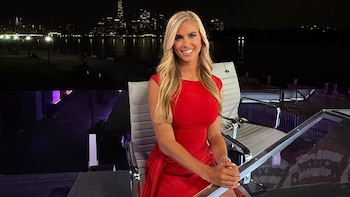 Una mujer rubia con vestido rojo sonríe sentada junto a una mesa transparente, con rascacielos iluminados de noche en el fondo