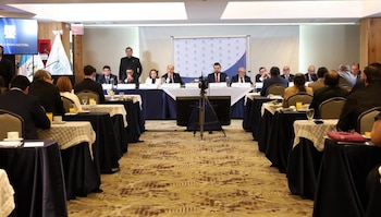 Vista frontal de una sala de conferencias con un panel de nueve personas sentadas en una mesa larga y numerosos asistentes en mesas redondas cubiertas