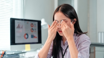 Una mujer con camisa a rayas se frota los ojos con ambas manos frente a una computadora en una oficina
