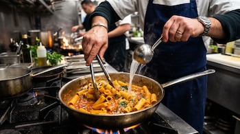 Manos de un cocinero que sostienen unas pinzas y un cucharón con líquido sobre una sartén con pasta y salsa de tomate en una cocina.