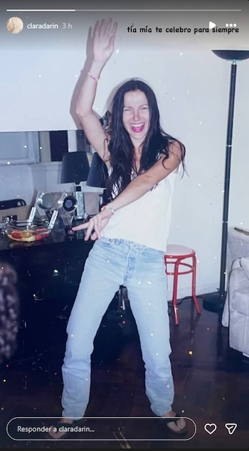 Mujer de pie con cabello oscuro, camiseta blanca y pantalón vaquero azul, tiene los brazos levantados y sonríe. Fondo con mesa, lámpara y un taburete rojo