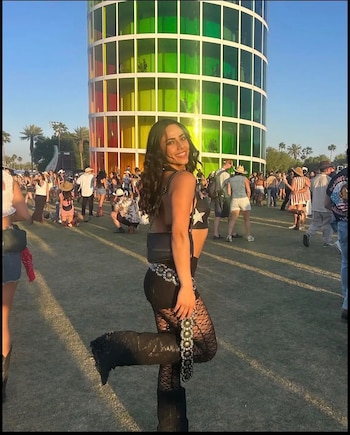 Una mujer joven con cabello oscuro sonríe, vestida con top negro y leggings de encaje, posando frente a una estructura cilíndrica de colores y una multitud en un festival