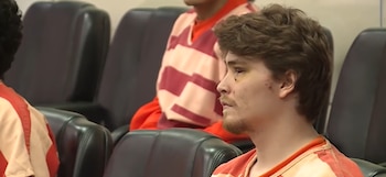 Hombre joven con cabello castaño, barba y marcas en su ojo izquierdo, vestido con uniforme naranja y rayas beige, sentado en una sala con otras personas al fondo