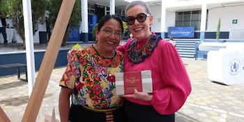 Dos mujeres posan en un patio exterior, una con huipil tradicional y otra con blusa fucsia y gafas, sosteniendo una caja de chocolate artesanal