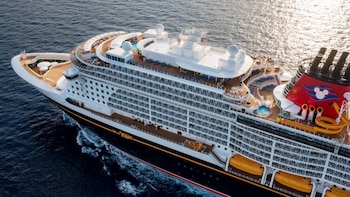 Vista aérea del crucero Disney Wish de Disney Cruise Line en el océano, con múltiples cubiertas, botes salvavidas naranjas y una chimenea roja con el logo de Mickey Mouse