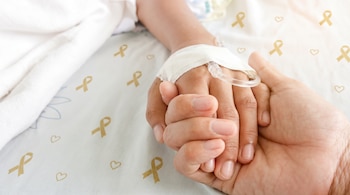 Primer plano de una mano adulta sosteniendo la mano de un niño con un catéter intravenoso, sobre una sábana blanca con dibujos de lazos dorados y corazones.