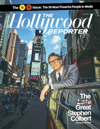 Stephen Colbert, un hombre de gafas en traje a cuadros, se asoma desde un autobús descapotable en Times Square, rodeado de edificios altos y pantallas publicitarias