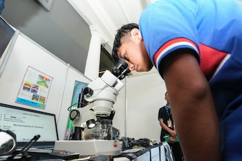 Un joven inclinado mira por un microscopio óptico frente a una mesa con rocas, frascos y una computadora portátil