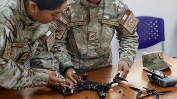 Dos personas con uniforme militar ensamblan un dron sobre una mesa de madera junto a herramientas, piezas y un control remoto