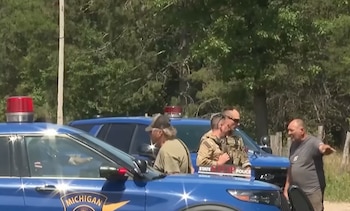 Dos vehículos de policía de Michigan, uno con luz roja en el techo, y cuatro hombres, algunos con uniforme táctico, junto a árboles y postes en un día soleado