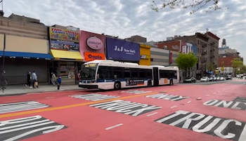 Un autobús articulado blanco transita por una avenida con carriles pintados de rojo y leyendas sobre el asfalto frente a varios comercios