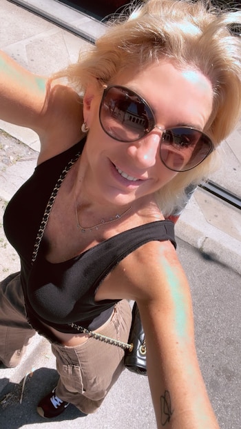 Mujer rubia de cabello suelto, con gafas de sol oscuras y un top negro, sonríe a la cámara en un día soleado al aire libre. Se ve parte de su brazo y hombro