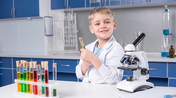 Un niño con bata blanca sostiene un tubo de ensayo junto a una gradilla con tubos coloridos y un microscopio sobre una mesa de laboratorio.