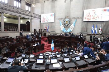 Interior de un salón legislativo con numerosos diputados sentados en sus escritorios con pantallas, varias banderas y el escudo nacional de Guatemala