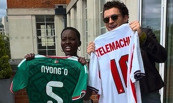 Lupita Nyong'o y Tom Holland sonríen mientras sujetan camisetas de fútbol. Nyong'o sostiene una verde de México; Holland, una blanca de Inglaterra