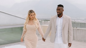 Un hombre con traje claro y una mujer con vestido beige caminan tomados de la mano sobre una azotea. Al fondo se observan montañas y un cielo gris.