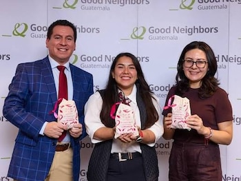 Un hombre y dos mujeres sonríen, cada uno sostiene una bolsa pequeña con una cinta rosa, frente a un muro con logos de Good Neighbors Guatemala