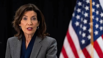 Retrato de la Gobernadora de Nueva York, Kathy Hochul, una mujer con cabello castaño oscuro, vistiendo un blazer gris, de pie frente a la bandera de Estados Unidos