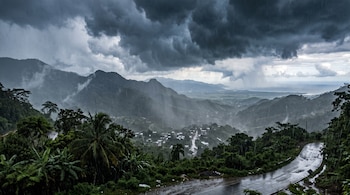 Nubes de tormenta y lluvia sobre un paisaje montañoso con una carretera asfáltica y un valle con casas.
