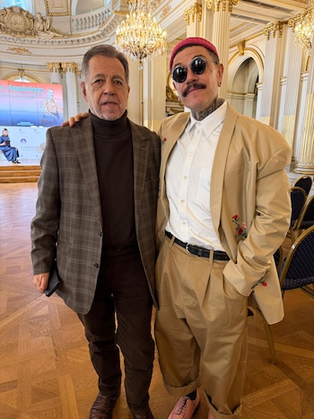 Dos hombres de pie en un salón elegante con paredes blancas y detalles dorados, un candelabro grande, y un podio con una pantalla al fondo