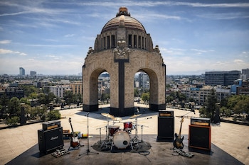 Escenario con batería, dos guitarras eléctricas, bajo y amplificadores frente al Monumento a la Revolución en la Ciudad de México, con vista urbana y cielo azul.