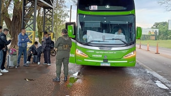 Un oficial uniformado de espalda, varias personas con equipaje, un autobús verde, una carretera, conos de tráfico y árboles