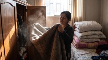Una mujer sostiene una frazada gruesa mientras el polvo se eleva de ella bajo un rayo de sol. Un armario abierto y una cama con edredones se ven al fondo.