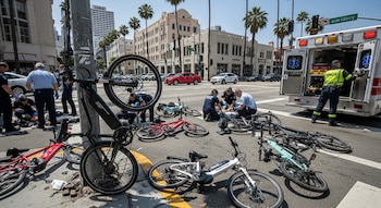 Una calle urbana con bicicletas eléctricas esparcidas y personal de emergencia atendiendo a varias personas en el suelo, con una ambulancia estacionada cerca.