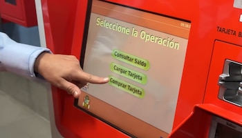Primer plano de una mano tocando la pantalla táctil de una máquina expendedora roja que muestra las opciones Consultar Saldo, Cargar Tarjeta y Comprar Tarjeta