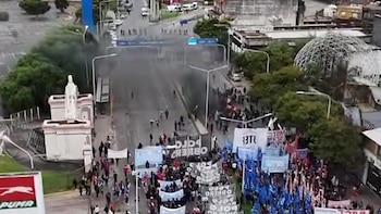 Vista aérea de una protesta masiva en una calle de Avellaneda, con cientos de personas, pancartas y banderas. Se observa humo en el centro, rodeado de árboles, edificios y una estatua blanca
