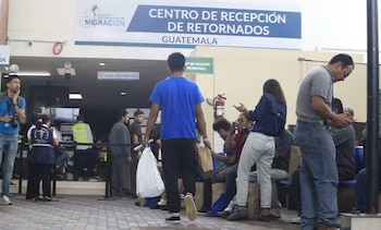 Un grupo de hombres sentados en sillas azules en una sala de espera utiliza teléfonos móviles y sostiene pertenencias personales