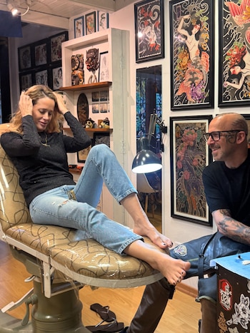 Una mujer sentada descalza en una camilla dentro de un local de tatuajes conversa con un tatuador con gafas y tatuajes en la cabeza