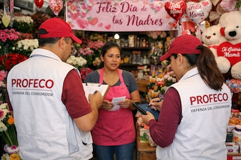 Dos inspectores de PROFECO, un hombre y una mujer, con gorras rojas y chalecos blancos, interactúan con una empleada en una florería.